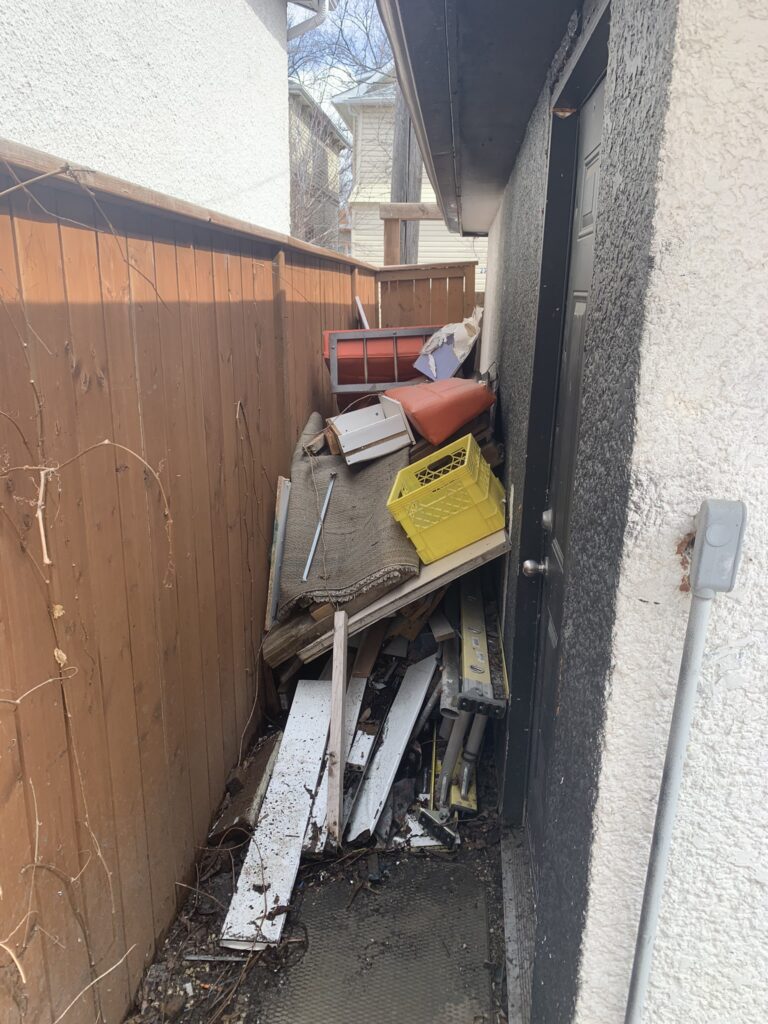 A nook between a garage and a fence that is littered with junk such as wood and furniture pieces