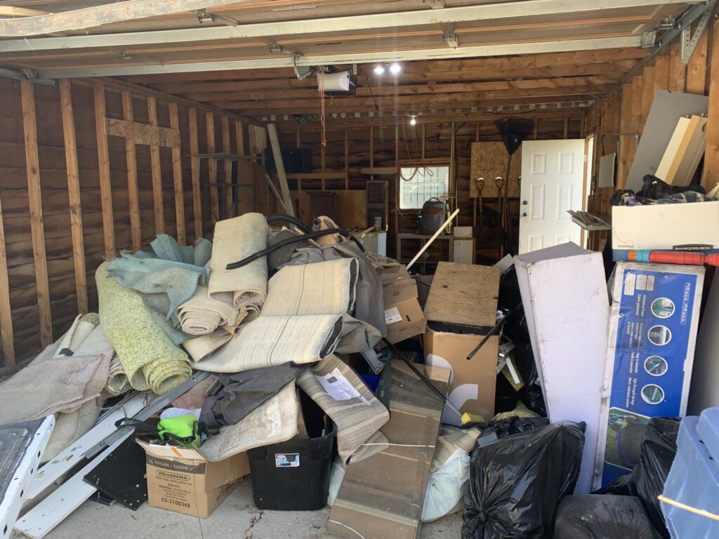 A garage that is filled with junk such as old carpets, boxes, wood, and shelves