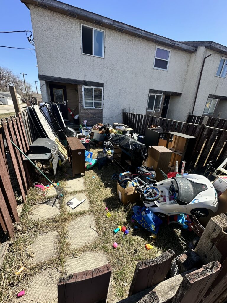 A back yard littered with junk such as children's toys, shelves, garbage bags, and mattresses